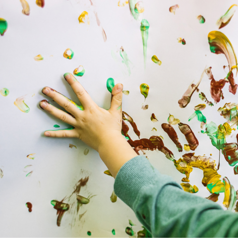 Kids hand finger painting with green, gold, and brown paint on white paper.