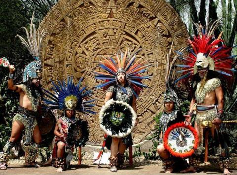 A group of dancers in traditional Aztec costume stand in front of a large calendar stone. 