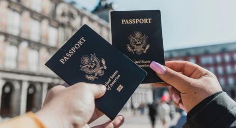 Two hands holding passport books with buildings in the background